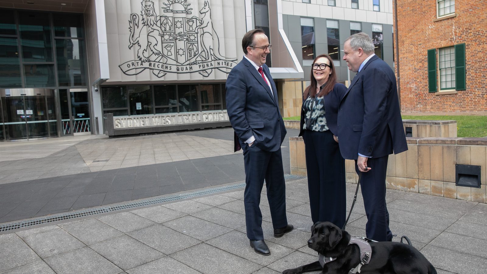 Timothy Roberts, Rebecca Grant and Vaughan Roles talking in the forecourt of the Sydney West Trial Courts, Parramatta, with Vaughan's guide dog Scooter lying at their feet.